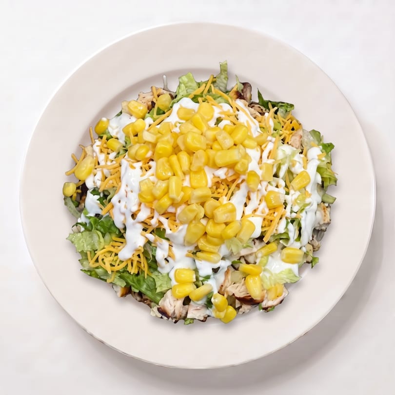 Chicken salad with lettuce, shredded cheese, corn, and white dressing in a white bowl.