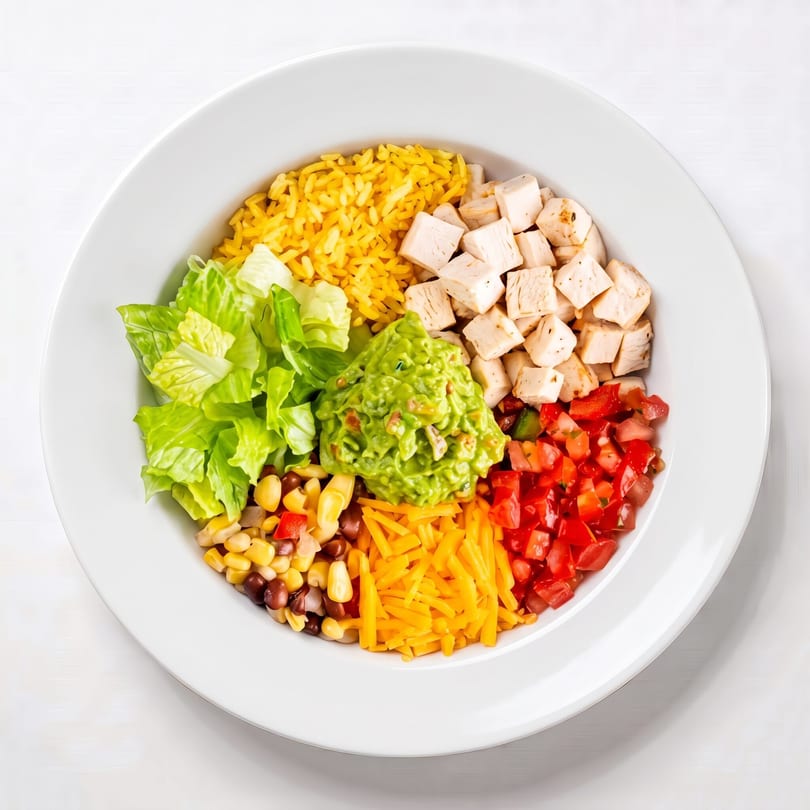Overhead view of a colorful chicken burrito bowl with rice, lettuce, guacamole, corn, beans, cheese, and tomatoes.