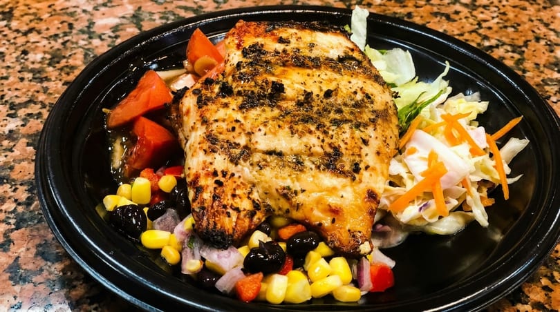 Grilled chicken breast with corn, black beans, tomatoes, and coleslaw in a black bowl.